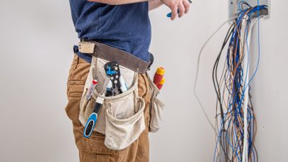 Electrician builder examines the cable connection in the electrical line in switchboard indoor. Electrician builder at work, examines the cable connection in the electrical line in the fuselage of an industrial switchboard. Professional in overalls with an electrician's tool.